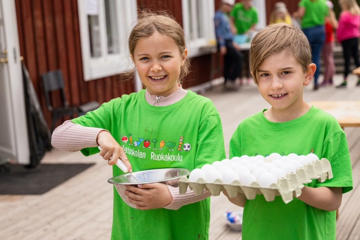 Kaksi lasta hymyilee vihreissä paidoissa. Toisella on kädessä kananmunia ja toisella kulho ja vispilä.