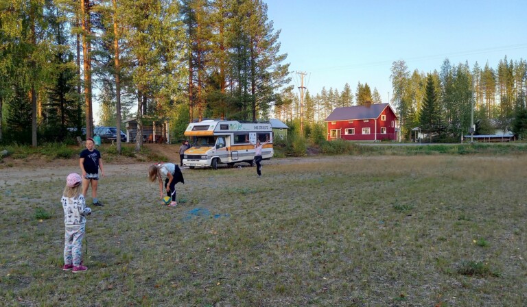 Kolme lasta kentällä. Taustalla matkailuauto ja maalaistalo.