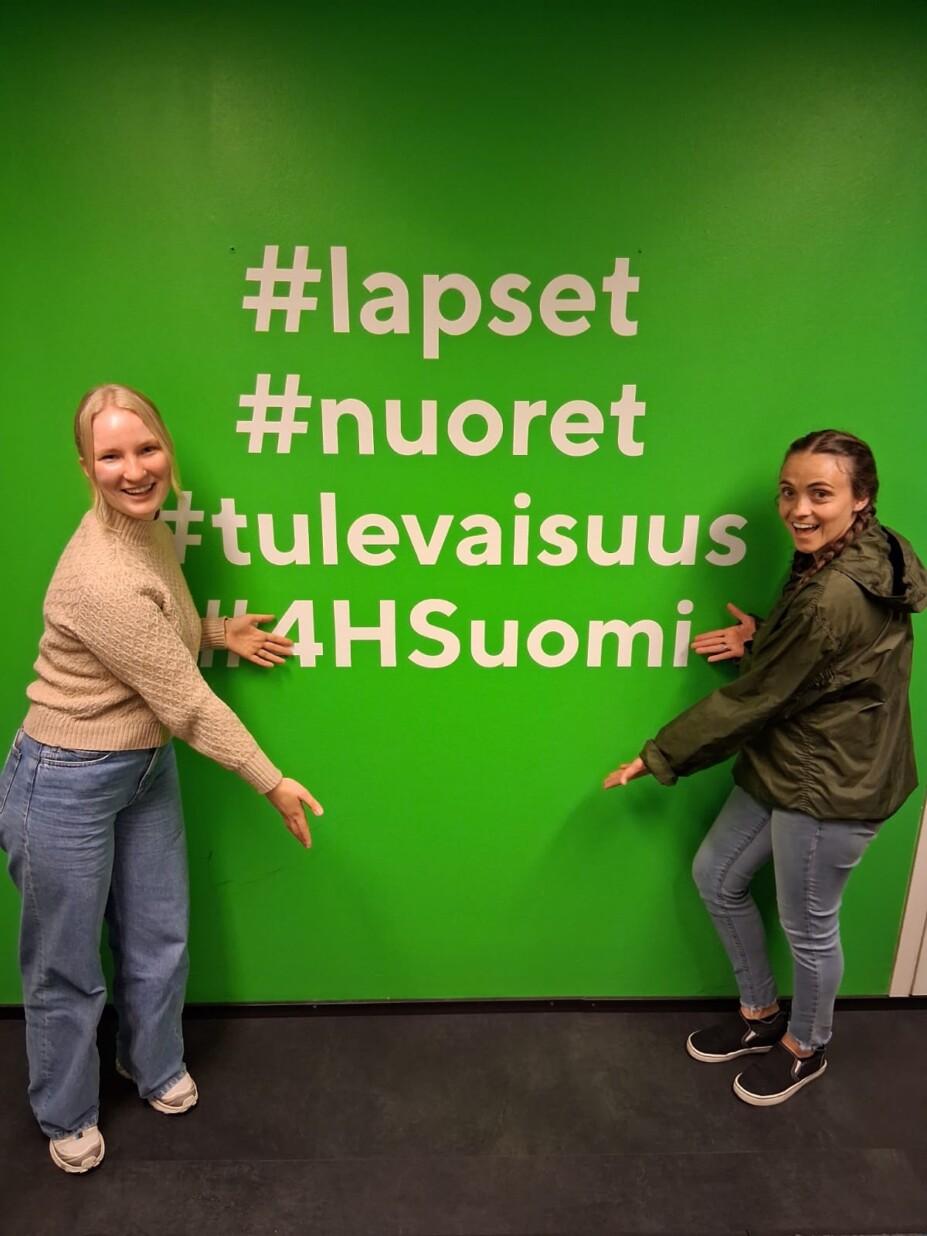Kaksi naista osoittaa seinällä olevaa tarraa, jossa lukee #lapset, #nuoret #tulevaisuus ja #4HSuomi.